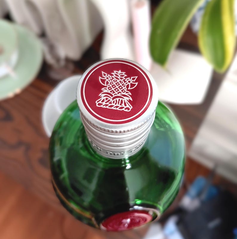 May include: A green glass bottle with a silver cap and a red top. The red top features a white pineapple design. The bottle is partially visible, showcasing its rounded shape and a red seal at the base.