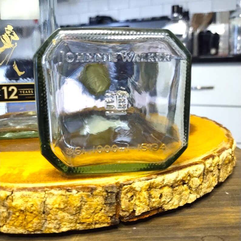 May include: A clear glass Johnnie Walker bottle with a green base, on a round, yellow-toned wooden slab. The bottle's label is partially visible, and the wood has a natural bark edge.