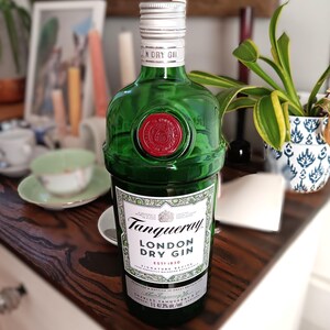 May include: A green glass bottle of Tanqueray London Dry Gin. The bottle has a silver cap and a red wax seal. The label features the brand name and product information. The bottle is displayed on a wooden surface.