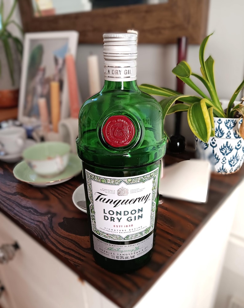 May include: A green glass bottle of Tanqueray London Dry Gin. The bottle has a silver cap and a red wax seal. The label features the brand name and product information. The bottle is displayed on a wooden surface.
