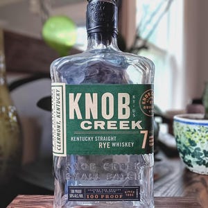 May include: A clear glass bottle of Knob Creek Kentucky Straight Rye Whiskey with a dark green label. The label features the text Knob Creek, 7, and 100 Proof. The bottle has a black cap and embossed text on the glass reading Knob Creek Small Batch.