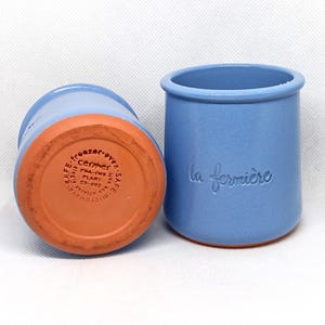 May include: Two blue ceramic crocks with a terracotta base. The crock on the left has the words "center raven SAFE FDA-IMS PLANT ES-002 service" printed on the base. The crock on the right has the words "la fermière" printed on the side.