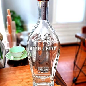 May include: A clear glass bottle of Angel's Envy Kentucky Straight Bourbon Whiskey finished in port wine barrels. The bottle features a tall, slender neck with a wooden stopper and a label displaying angel wings and the brand name in white text.