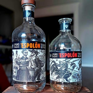 May include: Two clear glass tequila bottles with black labels featuring detailed skeleton illustrations. The taller bottle has a wooden cap and the label reads ESPOLON TEQUILA REPOSADO. The shorter bottle label reads ESPOLON TEQUILA BLANCO.