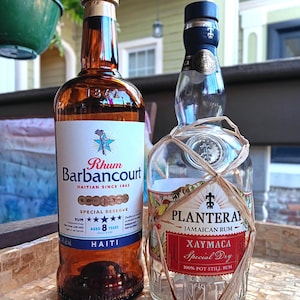 May include: Two glass bottles of rum. The left bottle is amber-colored with a white label reading Rhum Barbancourt, Haitian Since 1862, Special Reserve, Aged 8 Years. The right bottle is clear, wrapped in raffia, labeled Planteray Jamaican Rum Xaymaca.
