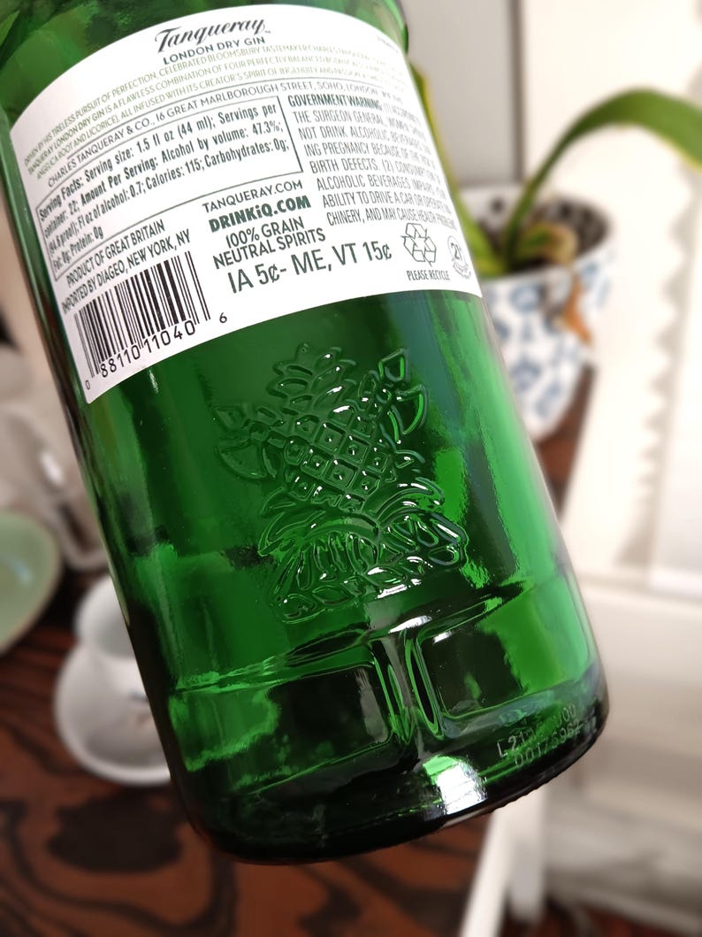 May include: A green glass bottle of Tanqueray London Dry Gin. The bottle has a pineapple emblem embossed on the glass. The label includes text about the product, including alcohol content and serving size. The bottle is partially visible.