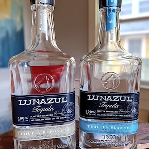 May include: Two clear glass bottles of Lunazul Tequila. One bottle has a silver label with "Tequila Reposado" and the other has a blue label with "Tequila Blanco". Both bottles have black caps and blue and silver labels.