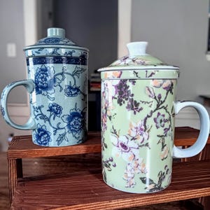 May include: Two ceramic tea mugs with lids. The left mug is light blue with a dark blue floral pattern. The right mug is light green with a multi-colored floral pattern featuring white, orange, and purple flowers. Both mugs have handles.