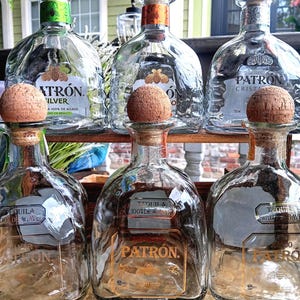 May include: Six clear glass Patrón tequila bottles arranged in two rows. The top row features Silver, Reposado, and Cristalino labels with cork stoppers. The bottom row shows three empty bottles with cork stoppers and gold-toned brand lettering.