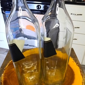 May include: Two clear glass bottles with black and white labels, set on a wooden, circular base. The bottles have a rectangular shape with angled sides. The background includes a kitchen stove and cabinets.