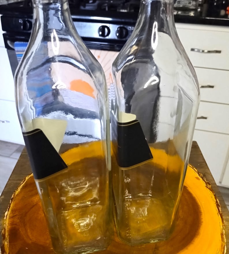 May include: Two clear glass bottles with black and white labels, set on a wooden, circular base. The bottles have a rectangular shape with angled sides. The background includes a kitchen stove and cabinets.