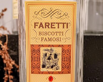 May include: A clear glass bottle of Faretti Biscotti Famosi liqueur. The label is primarily tan and gold with the brand name and a small illustration of a person near an oven. The bottle contains 750 mL and is 28% alcohol by volume.