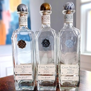 May include: Three clear glass tequila bottles with silver, gold, and gray stoppers. Each bottle has a label that reads "Casa Noble Tequila" and the type of tequila: Añejo, Reposado, and Blanco. The bottles are on a wooden surface.