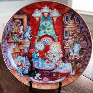 May include: Decorative plate featuring a whimsical scene with anthropomorphic cats in a Victorian-style interior. The plate has a gold rim and depicts cats holding hearts and enjoying tea. The background includes ornate furniture, curtains, and artwork.