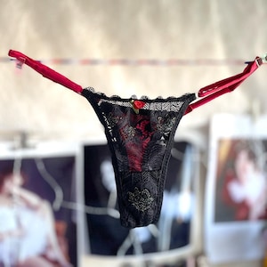 May include: Black lace thong with red straps and lining, featuring a small red rose. The lingerie is suspended from a clothesline, highlighting its delicate design. The image is set against a blurred background.