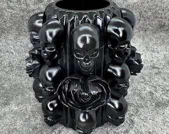 May include: A black ceramic vase with a skull and rose design. The vase is decorated with multiple skulls and roses around the exterior.