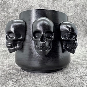May include: Black ceramic mug with three black skull-shaped handles. The mug has a textured, matte finish.