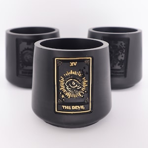 May include: Three black ceramic cups with gold accents. The centre cup features a tarot card design with the text "XV THE DEVIL".