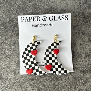 May include: Checkerboard crescent moon earrings with red heart accents. The earrings are handmade and have gold hooks. The earrings are displayed on a white card with the text "PAPER & GLASS Handmade".
