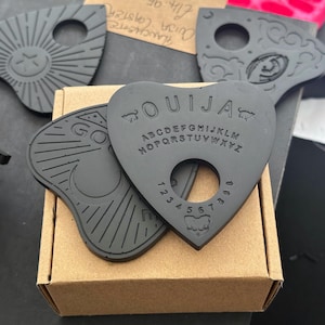 May include: Two black plastic Ouija boards. One board has the letters of the alphabet, numbers 1-9, and the words "yes", "no", and "goodbye". The other board has a sun and moon design.