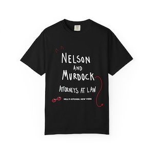 May include: A black short-sleeved t-shirt featuring white text that reads Nelson and Murdock Attorneys at Law and Hell's Kitchen, New York. The design includes a small red devil tail, red devil horns, and a pair of red circular glasses.