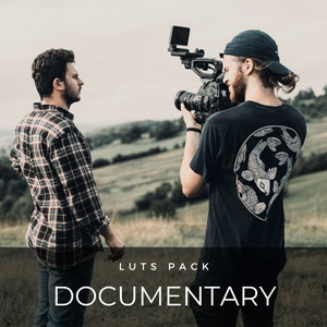 Puede incluir: Dos personas están filmando un documental. Una persona sostiene una cámara y la otra está de pie frente a la cámara. El fondo es un campo de hierba con colinas en la distancia.