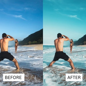 May include: A person taking a photo of another person standing in the ocean on a sandy beach. The person taking the photo is wearing black shorts and the person being photographed is wearing a black bikini.