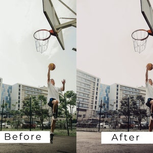 Op de afbeelding: Een persoon in een wit shirt en zwarte broek springt om een basketbal in een basket te schieten op een buitenbasketbalveld. Het veld is omgeven door gebouwen en bomen.