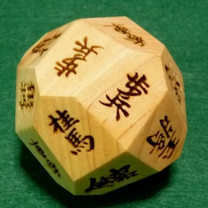 Peut inclure: Un dé dodécaédrique en bois avec des caractères chinois gravés. Le bois de couleur claire contraste avec les symboles gravés foncés. Le dé repose sur une surface verte.