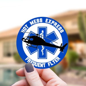 K&ouml;nnte beinhalten: Ein runder Aufkleber mit blauem Rand und wei&szlig;er Mitte, der ein blaues Stern-des-Lebens-Symbol, eine schwarze Hubschraubersilhouette sowie den Text "HOT MESS EXPRESS" und "FREQUENT FLYER" in blauen Gro&szlig;buchstaben am Rand zeigt.