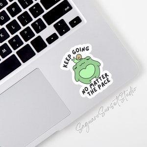 May include: A green frog with a snail on its head, a white sticker with the text "KEEP GOING NO MATTER THE PACE" on a silver laptop.