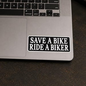 May include: A rectangular black sticker with a white border and white text that reads SAVE A BIKE RIDE A BIKER. The sticker is applied to the metallic surface of a laptop, positioned near the trackpad area, featuring a bold, sans-serif font.