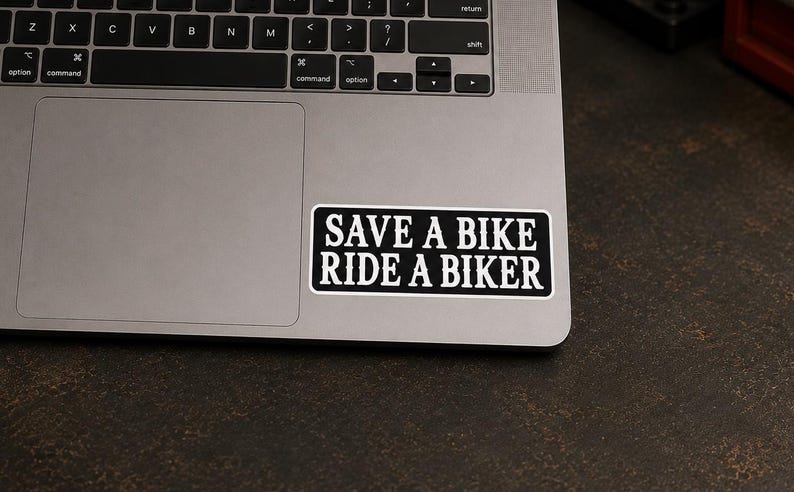 May include: A rectangular black sticker with a white border and white text that reads SAVE A BIKE RIDE A BIKER. The sticker is applied to the metallic surface of a laptop, positioned near the trackpad area, featuring a bold, sans-serif font.