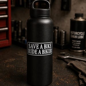 May include: A matte black insulated water bottle with a black screw-top lid featuring a carry handle. A rectangular white sticker is centered on the front of the bottle with black text that reads: SAVE A BIKE RIDE A BIKER in a bold, sans-serif font.