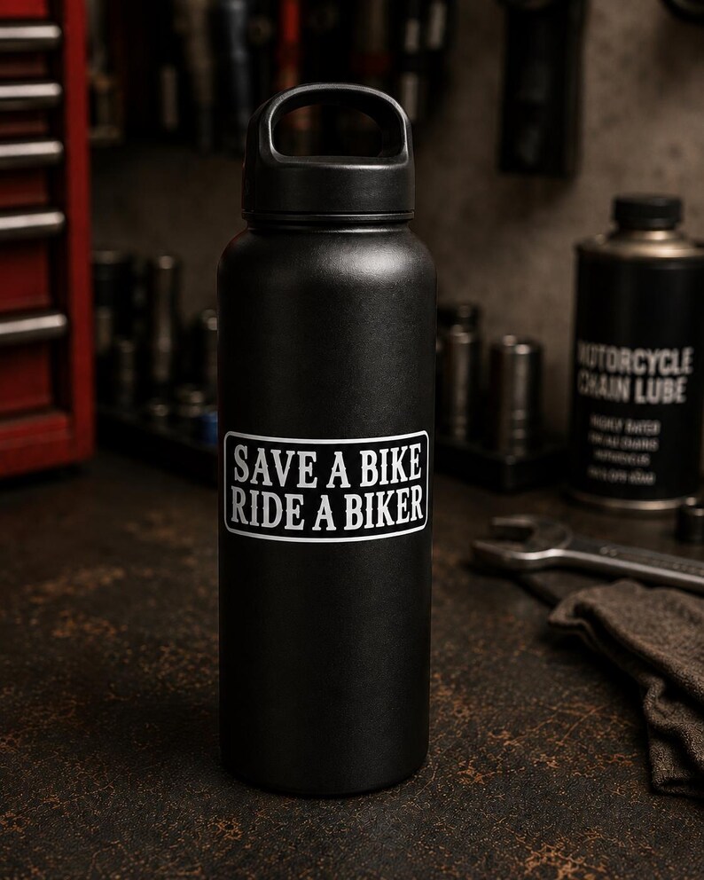 May include: A matte black insulated water bottle with a black screw-top lid featuring a carry handle. A rectangular white sticker is centered on the front of the bottle with black text that reads: SAVE A BIKE RIDE A BIKER in a bold, sans-serif font.