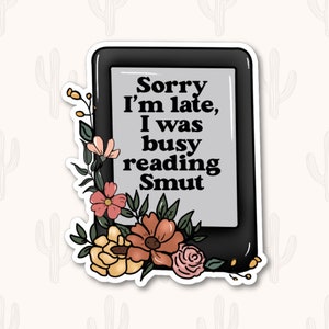 May include: A black and white sticker of an e-reader with the text "Sorry I'm late, I was busy reading Smut". The e-reader is surrounded by colorful flowers.