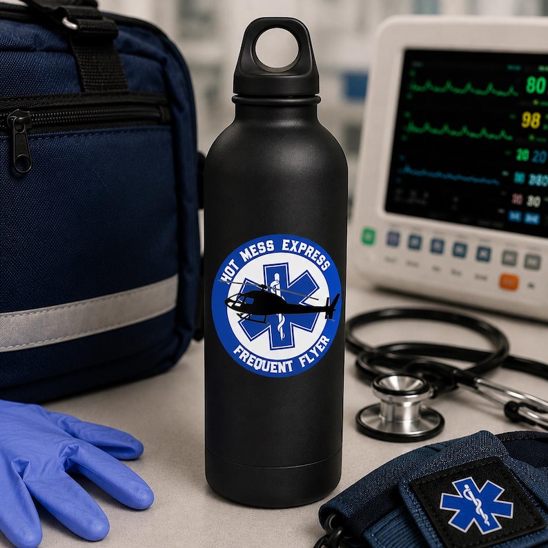 K&ouml;nnte beinhalten: Eine mattschwarze Wasserflasche mit einem runden Aufkleber, der ein blaues Star of Life-Emblem, eine schwarze Hubschraubersilhouette und den wei&szlig;en Text "HOT MESS EXPRESS FREQUENT FLYER" in einem kreisf&ouml;rmigen Rand um das Motiv zeigt.