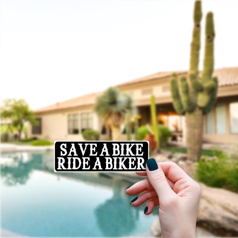 May include: A black and white rectangular sticker with the text "SAVE A BIKE RIDE A BIKER". The sticker is held in front of a blurred background of a house, pool, and desert plants.