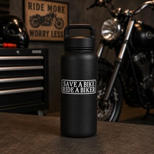 May include: A matte black insulated water bottle with a handle on the lid. The front of the bottle features a white rectangular sticker with black text that reads, "SAVE A BIKE RIDE A BIKER," printed in a bold, sans-serif, all-caps font style.