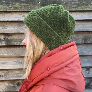 Puede incluir: Un gorro de lana verde con un borde doblado. El gorro lo lleva una persona con el pelo largo y rubio y una chaqueta roja.
