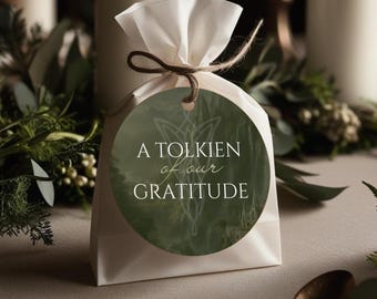 Puede incluir: Una bolsa de regalo blanca atada con hilo, con una etiqueta redonda que dice "A Tolkien of our Gratitude". La etiqueta tiene un fondo verde y una ilustración sutil. La bolsa está sobre una superficie neutra, con vegetación en el fondo.