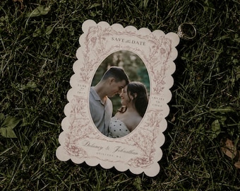May include: A rectangular Save the Date card with scalloped edges, featuring a central oval photograph of a couple, surrounded by intricate pink floral illustrations, with the text Delaney & Johnathan, June 27, 2017, and Quilcene, WA printed.