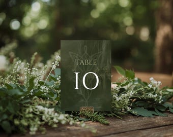 May include: A rectangular dark green table sign featuring the text TABLE 10 in white, centered above a white line-art emblem. The sign is held upright by a small metallic base, positioned amidst a display of white flowers and green foliage.