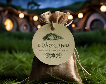 Puede incluir: Una pequeña bolsa de arpillera con una etiqueta redonda que dice "Thank You For Our Adventure!" en una fuente caprichosa. La etiqueta presenta una ilustración verde de un agujero de hobbit y detalles florales. La bolsa está atada con hilo y sobre césped verde.