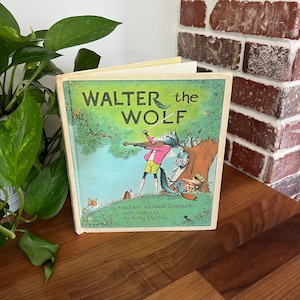 May include: A vintage children's book titled "Walter the Wolf" with a colorful illustration of a wolf playing the violin. The cover features a green background with a tree and other animals. The book is open on a wooden surface.