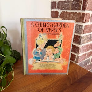 May include: Vintage children's book titled "A Child's Garden of Verses" by Robert Louis Stevenson, illustrated by Fern Bisel Peat. The cover features illustrations of children and floral elements, with text in a classic font. The book is on a wooden surface.