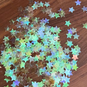 May include: A large quantity of iridescent star-shaped confetti in shades of green, blue, and purple. The confetti is scattered on a wooden surface.