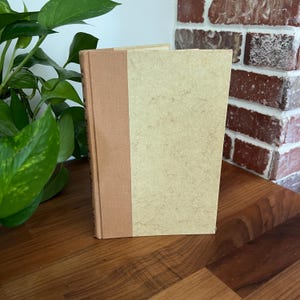 May include: A vintage book with a tan cover and a brown spine. The book is standing upright on a wooden surface, with a brick wall in the background. The book's pages are a light beige color.