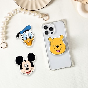 Puede incluir: Una funda transparente para teléfono con un soporte para teléfono de Winnie the Pooh amarillo y tres pegatinas de personajes de dibujos animados: Mickey Mouse, Pato Donald y Winnie the Pooh. Las pegatinas son coloridas y presentan las caras de los personajes.