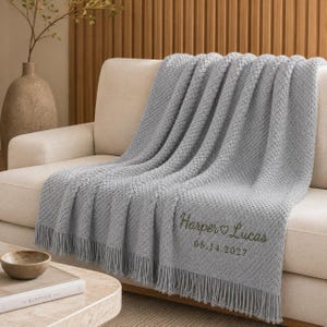 Puede incluir: Una manta tejida de color gris claro con un patrón de textura en relieve y flecos en los bordes. La manta presenta un bordado en hilo verde oscuro con el texto Harper ♡ Lucas y la fecha 14.06.2027 en la esquina inferior derecha.